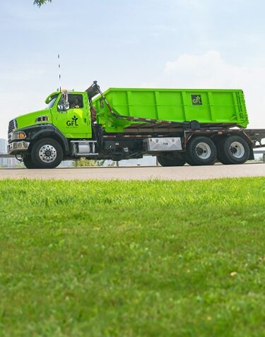 GFL truck transporting a dumpster on the road with green grass in the foreground