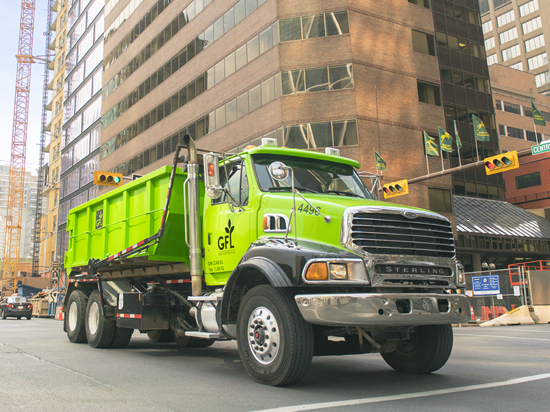 Green GFL truck transporting a dumpster in the middle of a downtown cityscape area