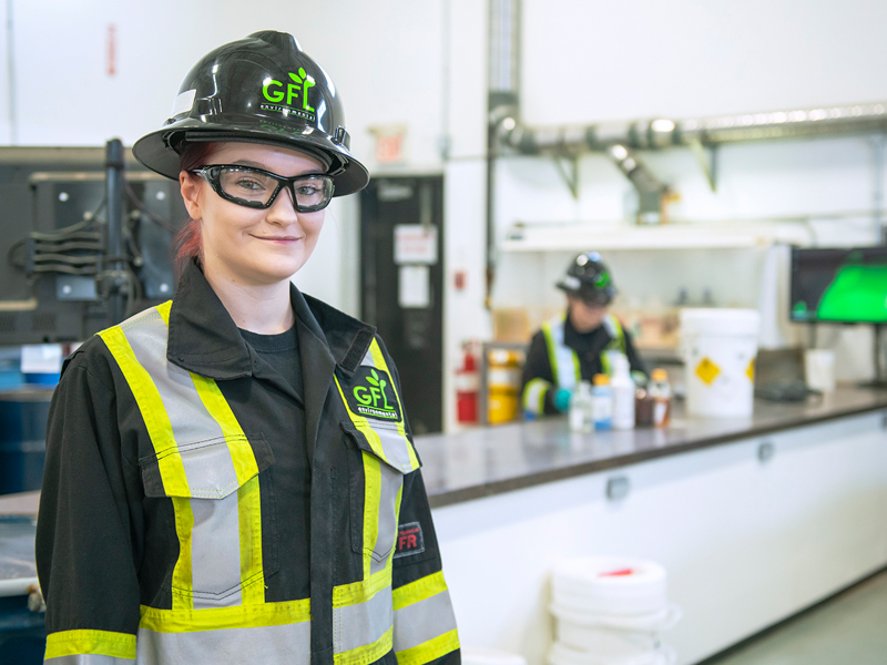 A GFL employee wearing goggles and a hard hat smiles while her coworker works in the background