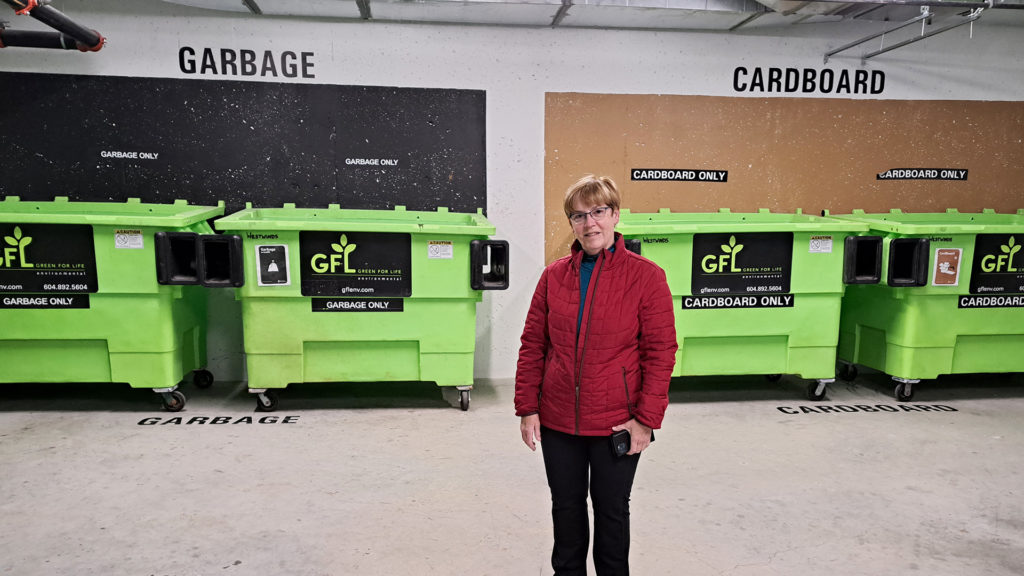 Pennie McNutt, employée de GFL, debout devant des bennes à ordures vertes de GFL marquées d’une affiche « poubelle » et « carton ».