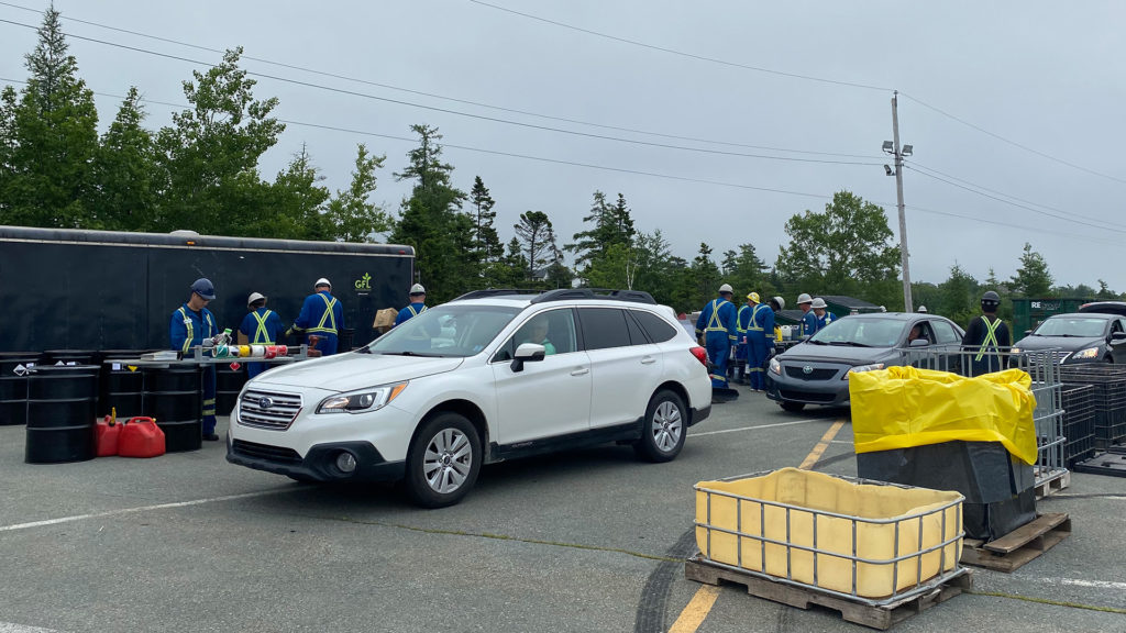 Cars lined up to drop off Household Hazardous Waste with GFL employees and a trailer in the background and bins to contain the different types of waste in the foreground. 